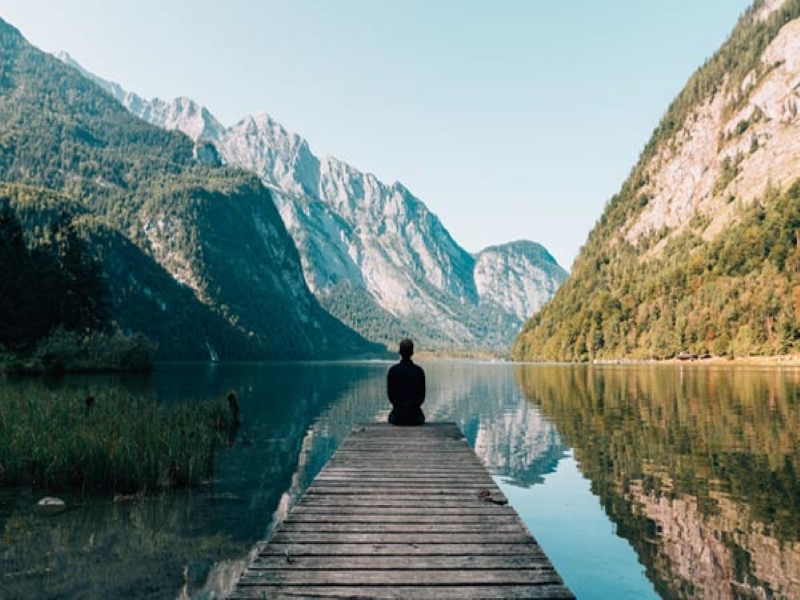 Un homme est assis sur un pontont face à un paysage de montagnes qui se reflète dans un lac